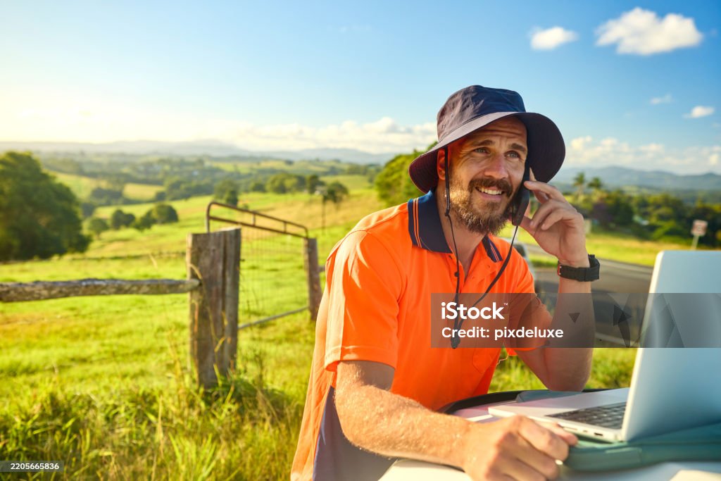 Smiling engineer wearing high-visibility shirt and hat using laptop and phone in a rural landscape, planning a construction project in australia