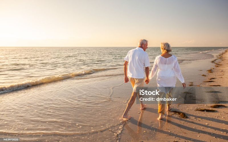 Happy senior man and woman old retired couple walking and holding hands on a beach at sunset