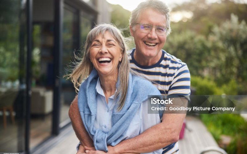 Mature couple standing arm in arm together and laughing outside in the back yard at home in summer