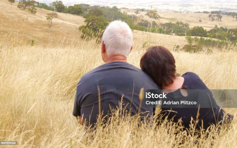 An elderly couple is sitting in the grass on top of a hill looking down into the valley. She is leaning against him
