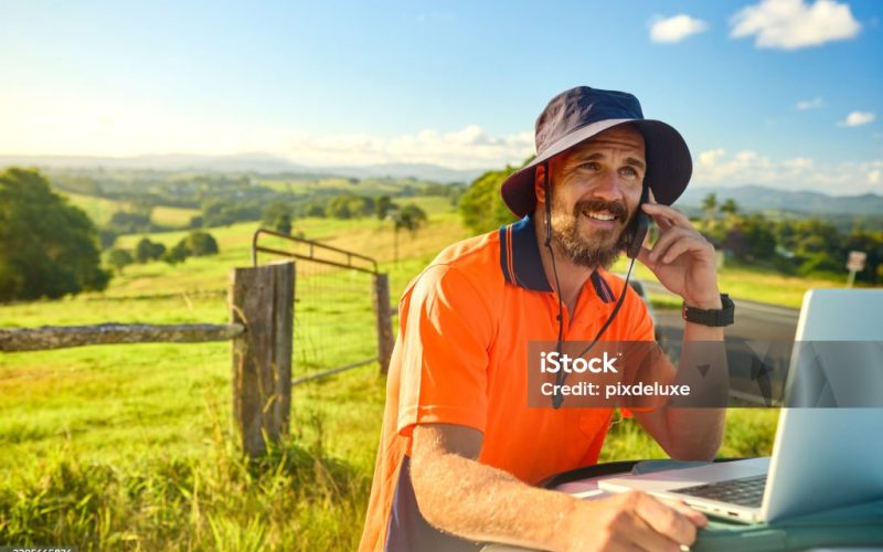 Smiling engineer wearing high-visibility shirt and hat using laptop and phone in a rural landscape, planning a construction project in australia