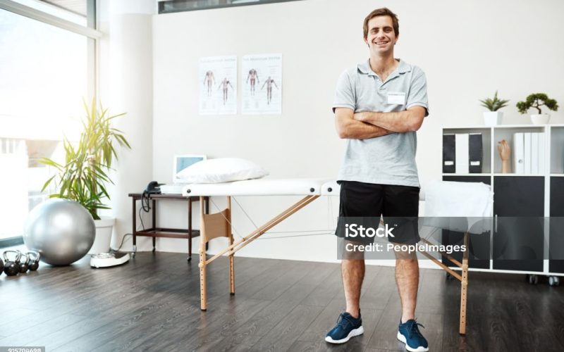 Portrait of a confident young male physiotherapist standing with his arms folded inside of his office