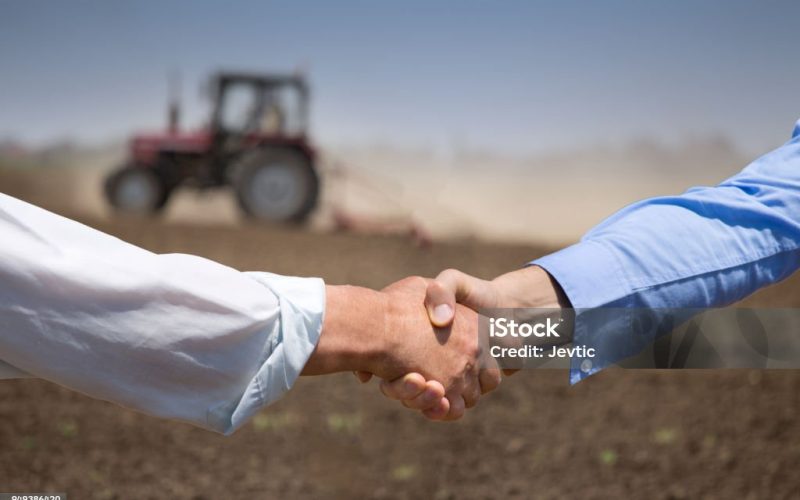 Two businessmen shaking hands in field with tractor working in background. Agribusiness concept